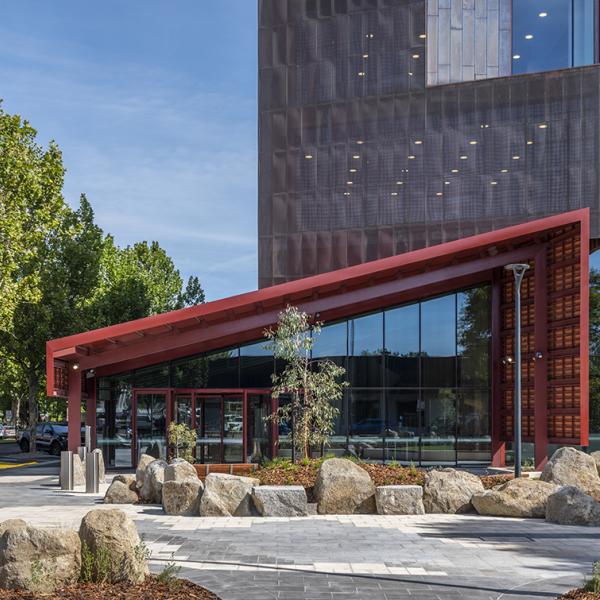 A paved and landscaped area looks towards a vaulted awning sitting over glass doors